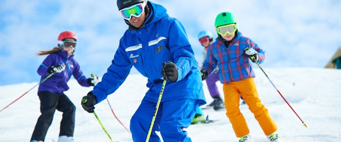 Picture of an adult on three children skiers dressed in colorful winter gear. They are skiing on a snowy hill.