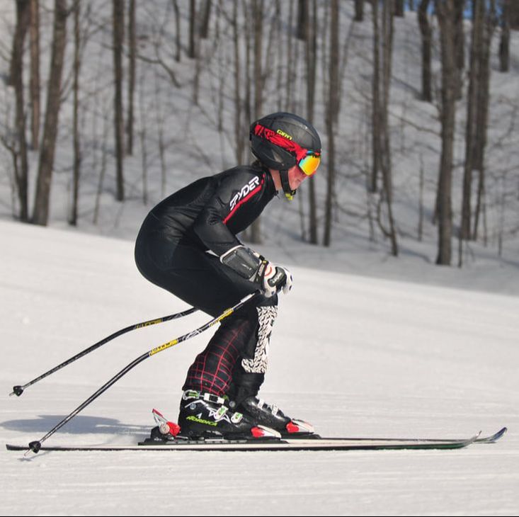 Picture of a ski racer dressed in a ski racing suit with a black helmet, goggles, and race skis. They are skiing on white snowy slopes and there are trees in the background. 
