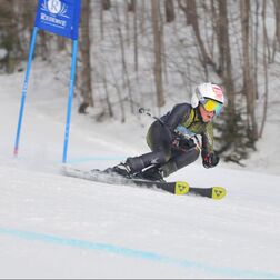 A young adult ski racer is racing down a snow hill. They are wearing a black and yellow ski racing suit, a white helmet, and skis, There are trees in the background.