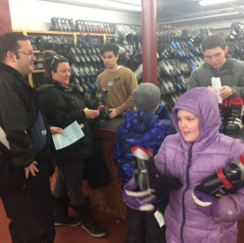 Picture of a family of four obtaining their ski boots from two employees in the rental area. The family is dressed in winter gear and are interacting with the employees who are asking what size boots they wear. In the background are racks of boots designed for skiing and snowboarding.