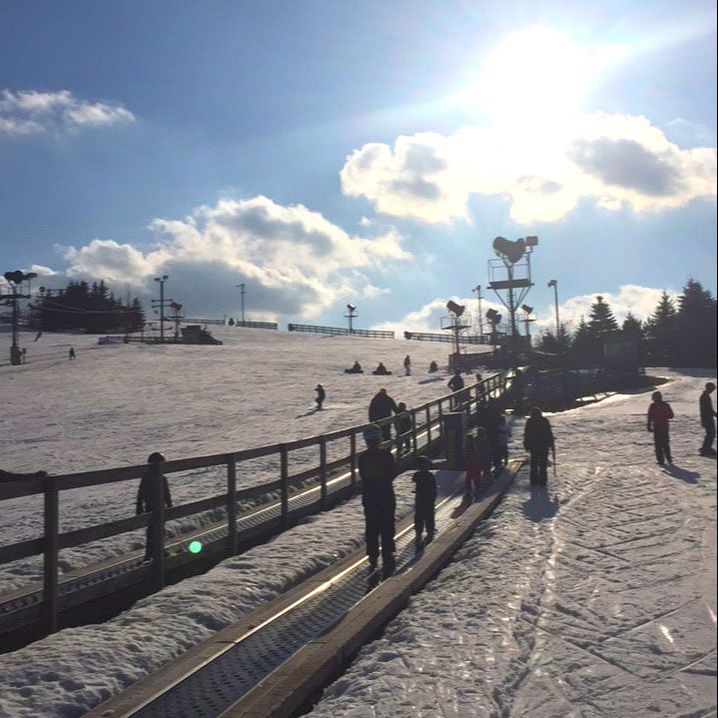 This image is of the lift in the beginners area at Pine Knob. New skiing and snowboarding students can step on to this lift which is essentially a conveyor belt and it gently carries them to the top of a very small hill where they can begin to learn. The picture was taken on a sunny day and there are clouds in the sky and there is snow on the ground.