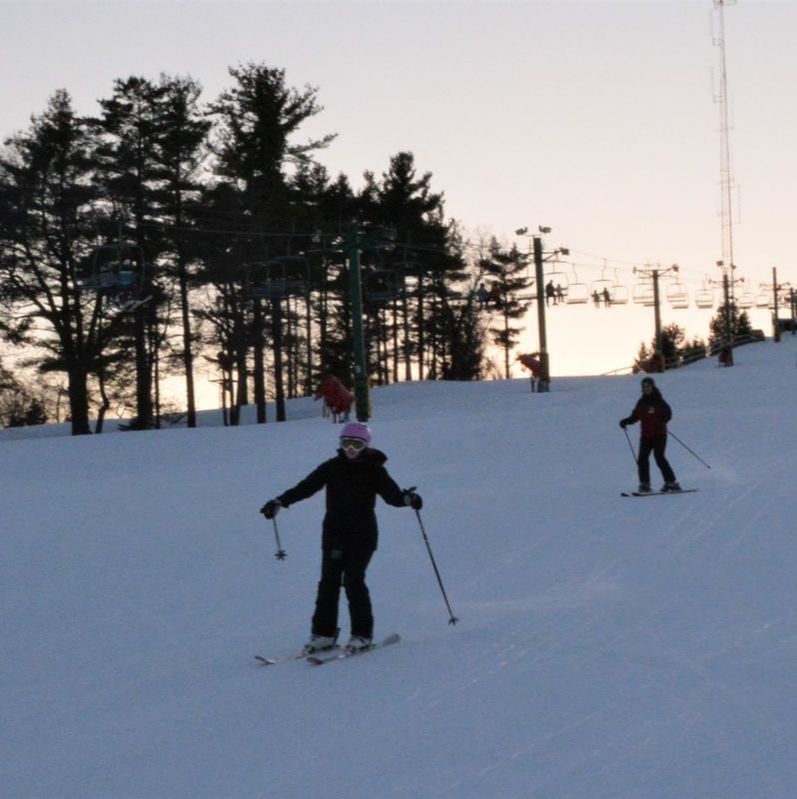 Picture of a private ski lesson at Pine Knob. A ski instructor in a red coat, black snow pants, and a helmet is skiing with her student coaching her. The student is a young woman wearing all black and a pink helmet. They are skiing on a snow hill and there are pine trees behind them.