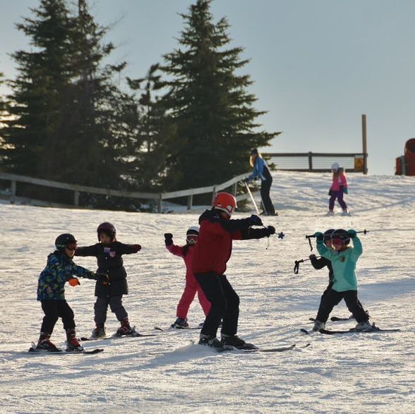 Picture of an adult Pine Knob ski instructor who is wearing a red coat, black ski pants, a red helmet, and he is in his skis on the snowy hill. His students are following his lead. They are four younger children who are learning to improve their turns as they ski down the hill. There are pine trees in the background.