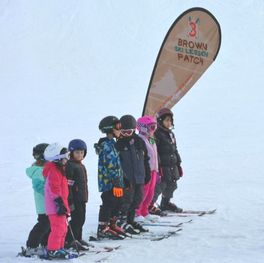 Picture of young students lined up and ready to take a skiing lesson. The children are dressed in bright winter gear including coats, snow pants, helmets, gloves, and skis. They will be spending an hour with their instructor out on the slopes.