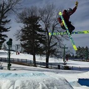 Picture of a skier doing a jump at the Pine Knob terrain park. There is snow and a blue sky,