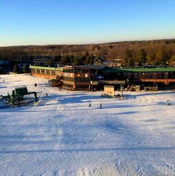 Picture taken from up on a hill looking down at the Pine Knob Resort Lodge. The building is large and has many windows. The sky is blue and there are many trees behind the building.