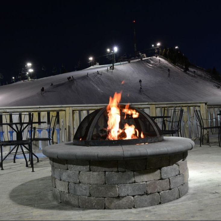 Picture of the outdoor warming fire on the upper deck of the lodge at Pine Knob. In this image the fire is surrounded by tables and chairs. They overlook the ski hill which is covered in snow and the sky is a dark because it is nightime and the lights are turned on all over the hill.