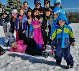Picture of four adult Pine Knob Ski School instructors and eleven children who are students. Everyone is dressed in colorful winter coats, snow pants, and gloves. They are outdoors on a sunny day, standing on the snow and smiling at the camera.
