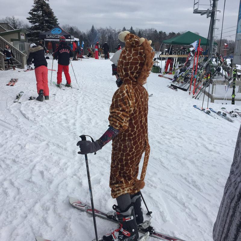 Picture of a child wearing a giraffe costume and enjoying an event at Pine Knob. There are other people outside and everyone is wearing skis. The ground is covered in snow and there is a tent with skis that people can try out.
