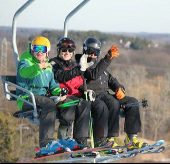 Picture of three men riding the chair lift at Pine Knob, They are wearing winter gear including coats, helmets, snow pants, and gloves. They are all wearing their skis and are riding the chairlift to the top of the hill so that they may ski down.
