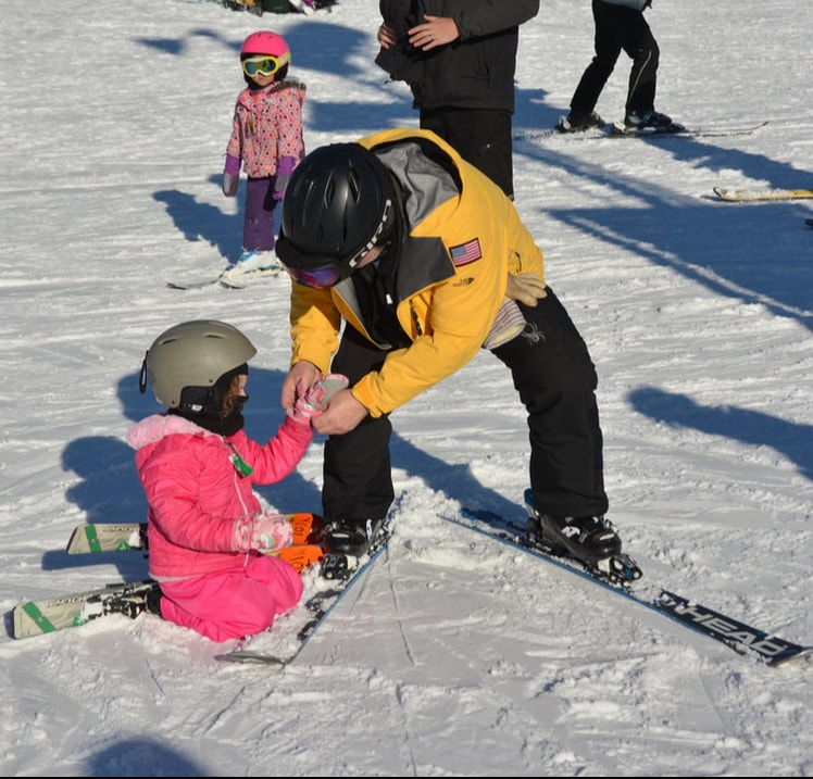 Picture of Pine Knob Ski School instructor Mike. He is in a red ski instructor coat and snow pants. He is helping a young child with the placement of his skis and they are outdoors enjoying the snow.