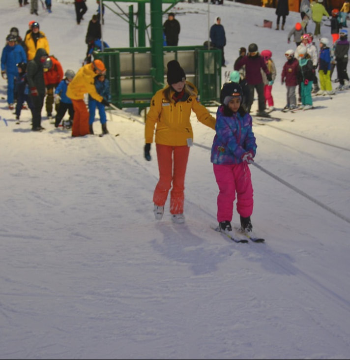 This image is a family of four who are walking from the lodge at Pine Knob out to the beginner area to start their first lesson. They are wearing winter clothing, are carrying skis, and are smiling at the camera.