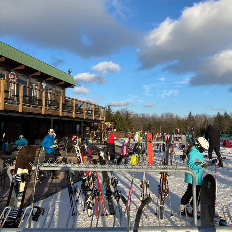 Picture of the second floor deck at Pine Knob that looks out on the resort. The sky is a deep blue and the snow is bright white. The deck has tables and chairs that guests enjoy sitting in.
