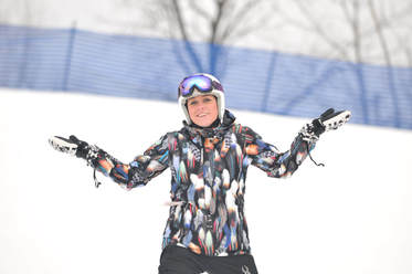 Picture of an adult woman who is wearing a patterned ski coat, snow pants, and a white helmet. She is outdoors, standing on the snow and she is facing the camera, smiling and has her hands raised up to shoulder height.