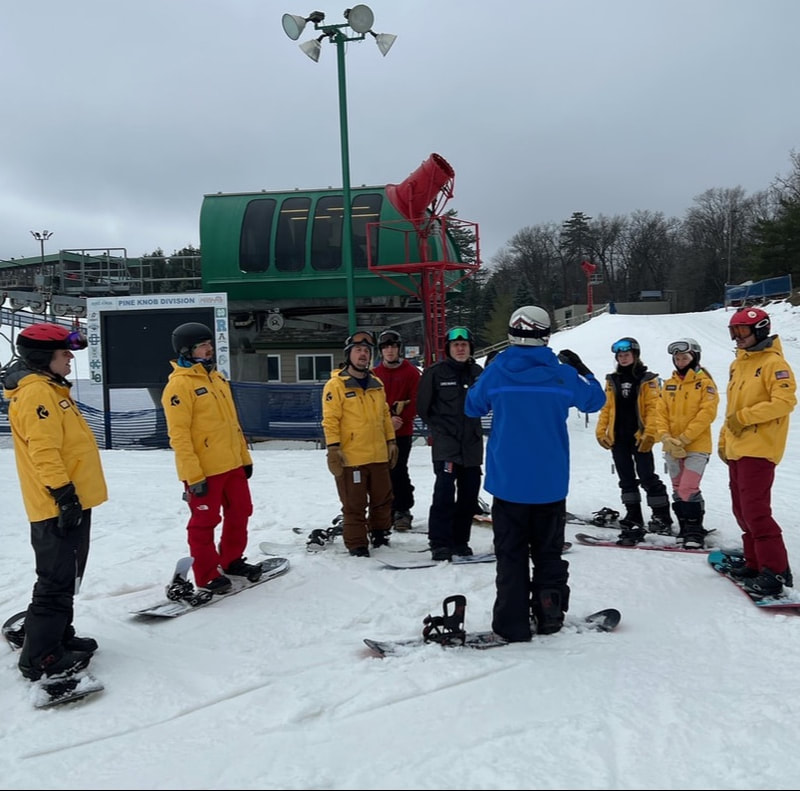 Picture of a Pine Knob ski instructor wearing a red coat and black helmet. He is teaching a student wearing an orange coat and black helmet. They are on the snowy slopes of Pine Knob and are wearing skis.