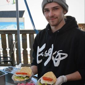 Picture of a young man smiling. He is standing on the deck that is off the second floor of the Pine Knob lodge. He is holding fresh cooked food from the grill and smiling at the camera.