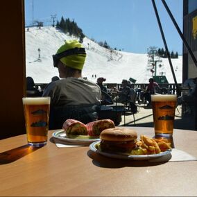 Picture of a burger, french fries, and beer sitting on the table in the cafeteria at Pine Knob. It is next to a large window that is looking outside towards the large ski hill covered in snow. A guest is sitting outside just on the other side of the window in one of the chairs that are near the firepit area on the deck,