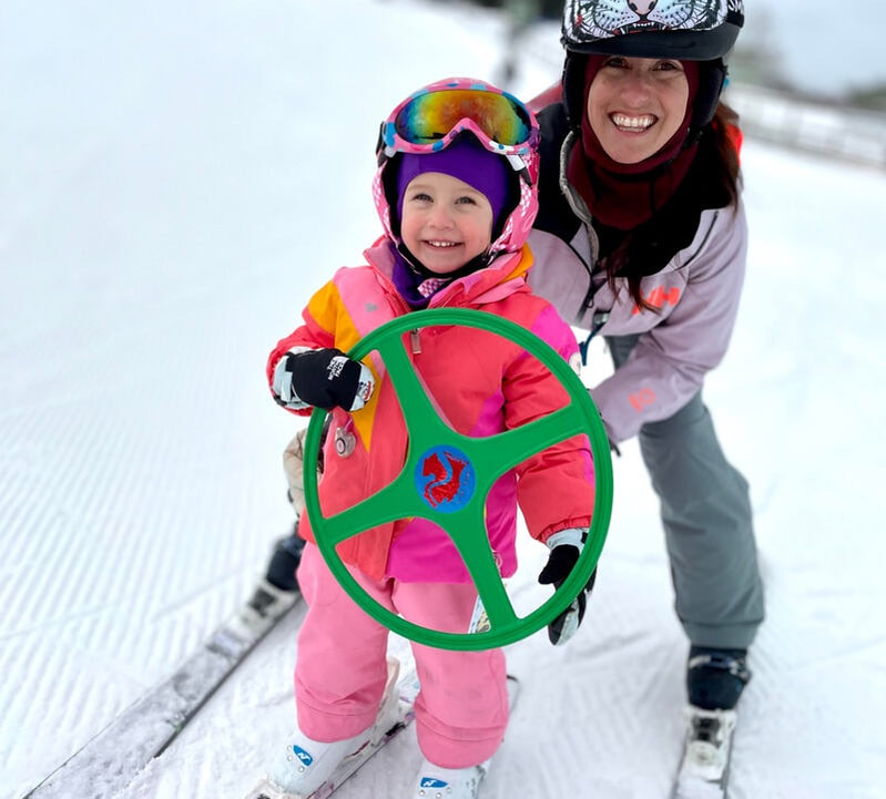 Picture of an adult woman and three children dressed up in colorful ski gear, helmets, and ski goggles, smiling at the camera. Text in this photo states 