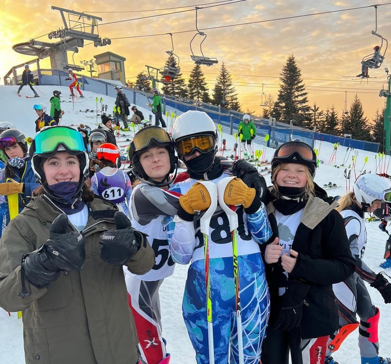 Picture of Coach Tom Lining and his ski racing students. Everyone is outdoor on the snowy hill and bundled up in colorful winter gear including coats, snow pants, helmets, and goggles at Pine Knob.