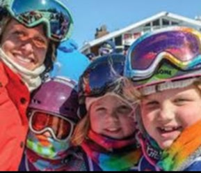 Picture of an adult woman and three children dressed up in colorful ski gear, helmets, and ski goggles, smiling at the camera. Text in this photo states 