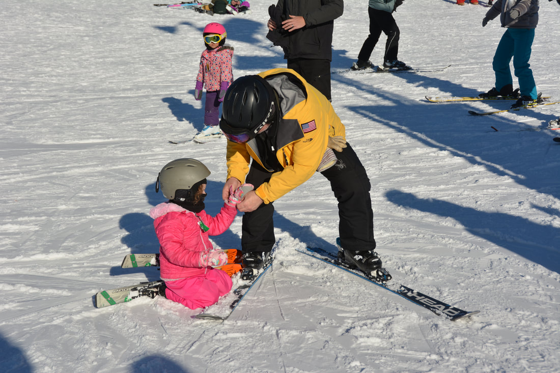 This picture is of an adult snowboarding instructor wearing a purple coat and black ski pants. They are leading a group of children who are learning to snowboard. There are six children in the class wearing colorful coats and winter gear. They are following the instructor with their snowboards on the snow covered hill at Pine Knob.